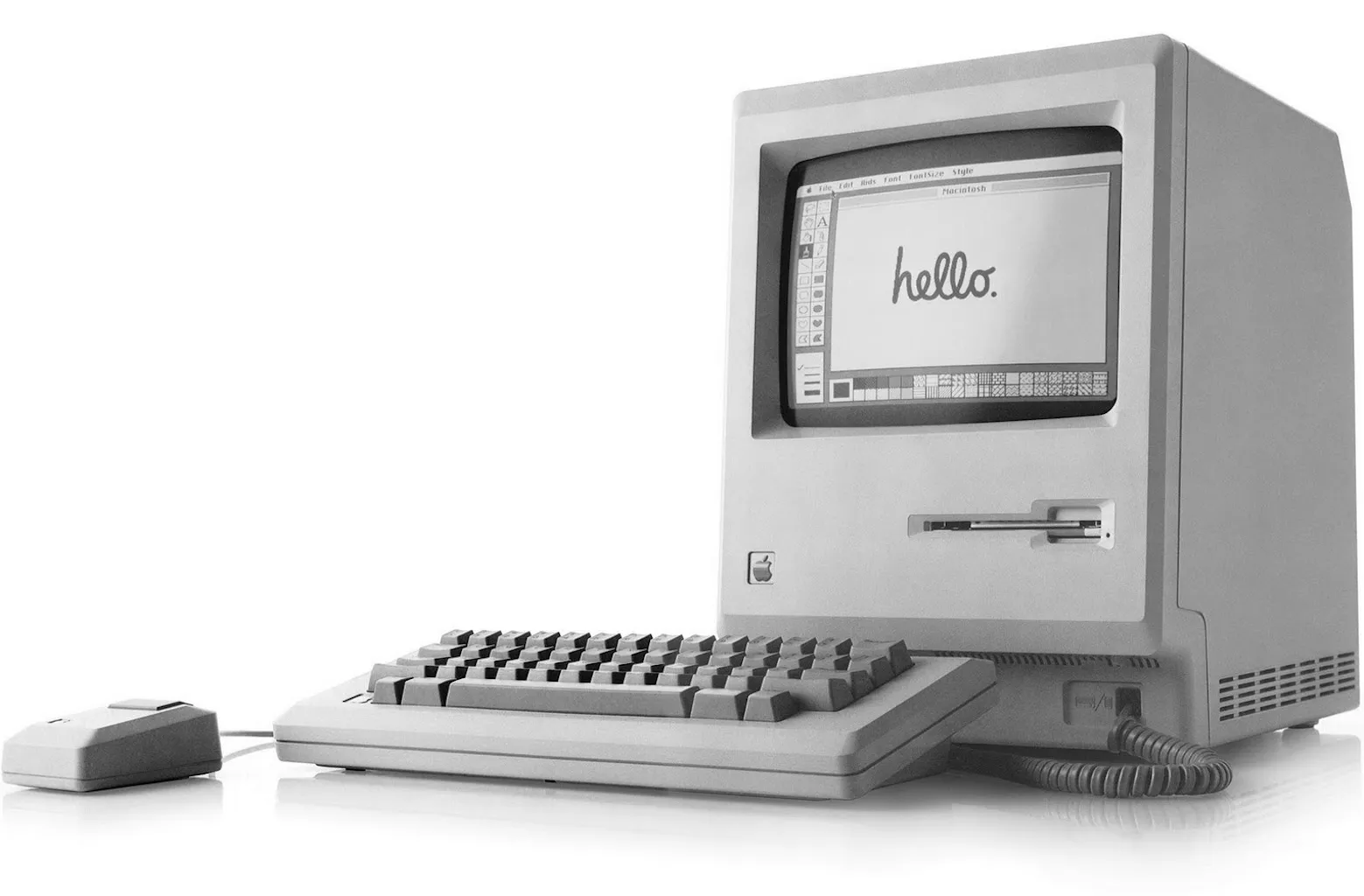 The Macintosh 128K, a beige box with a small screen and a floppy disk drive, sits beside a keyboard and mouse. The screen displays the classic 'hello' motif used frequently in Apple’s advertising.