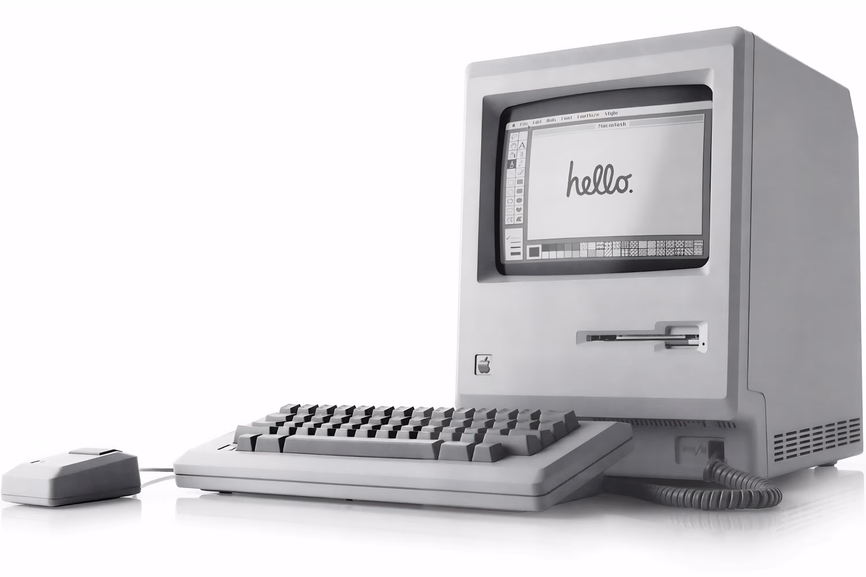 The Macintosh 128K, a beige box with a small screen and a floppy disk drive, sits beside a keyboard and mouse. The screen displays the classic 'hello' motif used frequently in Apple’s advertising.