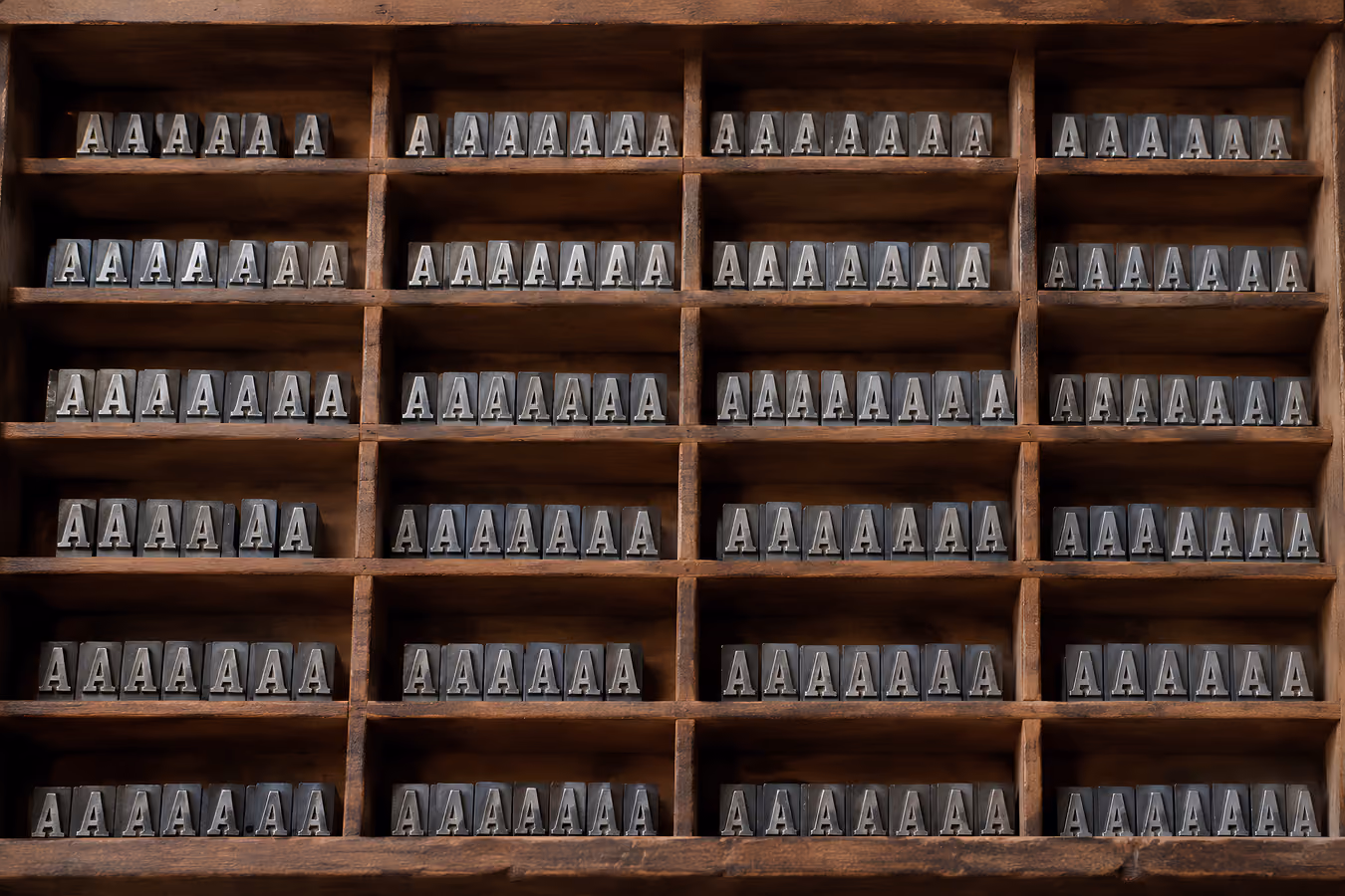A wooden printer’s type case divided into rows of compartments, each filled with identical metal ‘A’ letters lined up side by side.